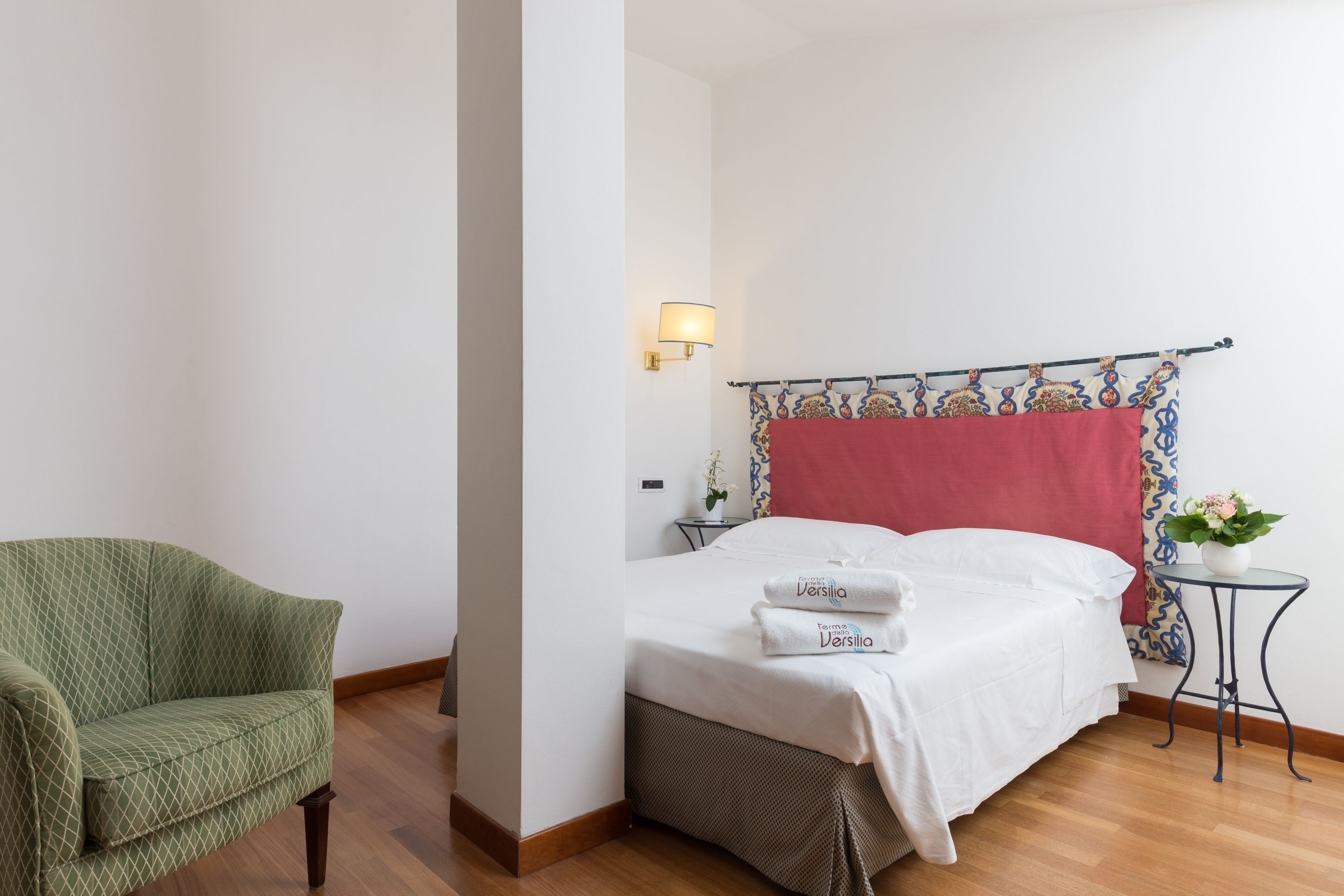 Een netjes ingerichte hotelkamer met een bed, een groene fauteuil, een nachtkastje met een lamp en een kleine tafel met een plant.