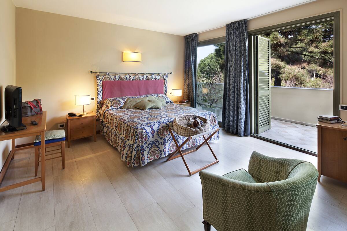 Una camera da letto con un letto, comodini, una scrivania, una sedia e un balcone affacciato sugli alberi.