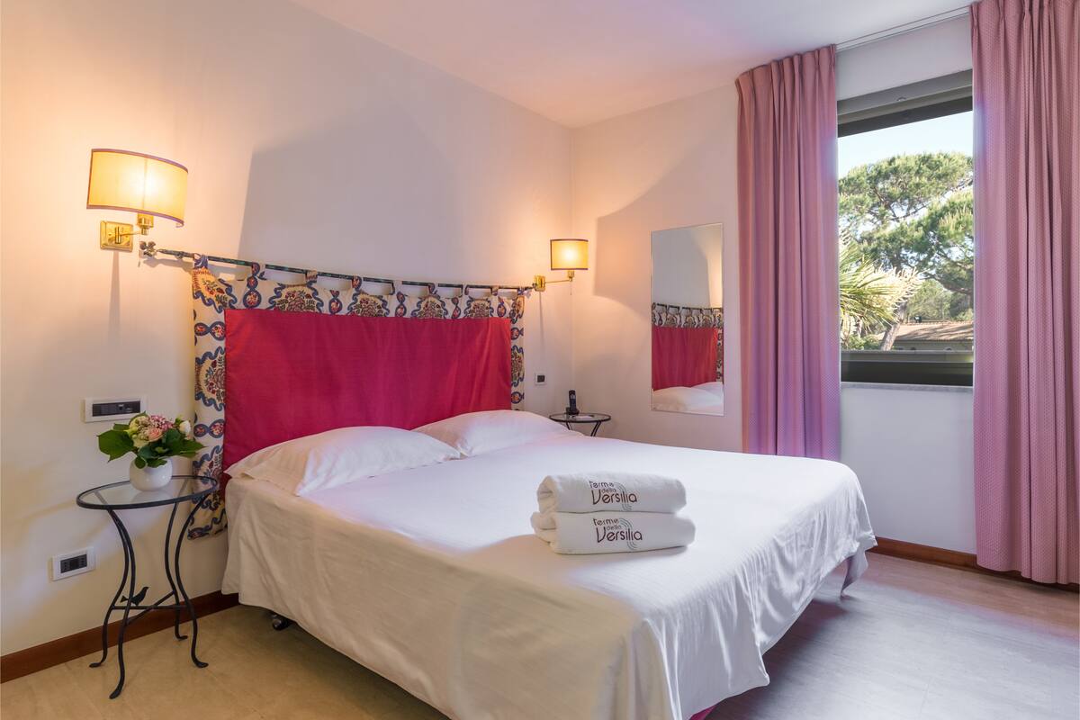 Una camera d'albergo con un letto, due lampade ai lati, uno specchio, un tavolino con un vaso di fiori e una finestra con tende.