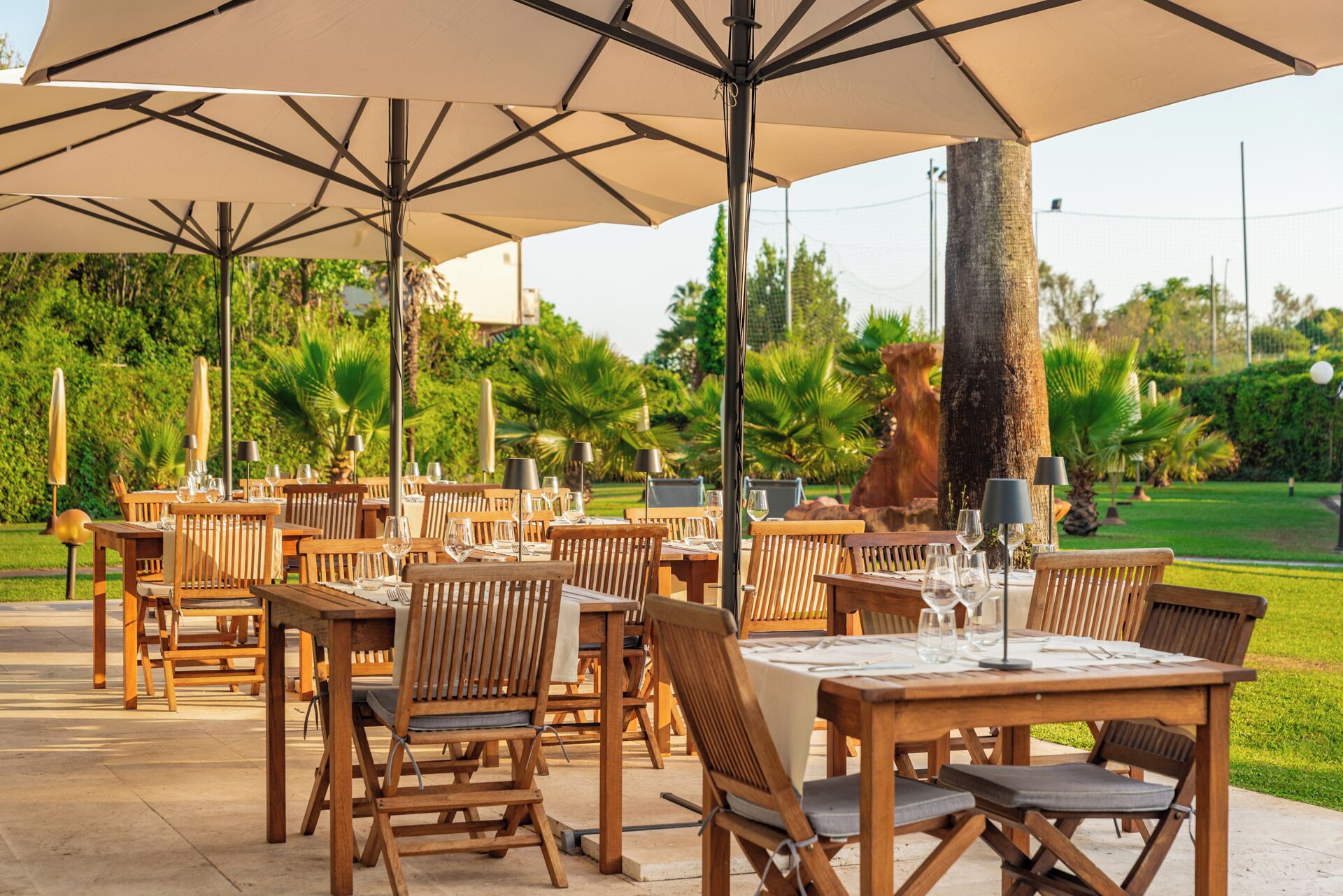 Un área de comedor al aire libre con mesas y sillas de madera, sombrillas grandes y un jardín bien cuidado al fondo.