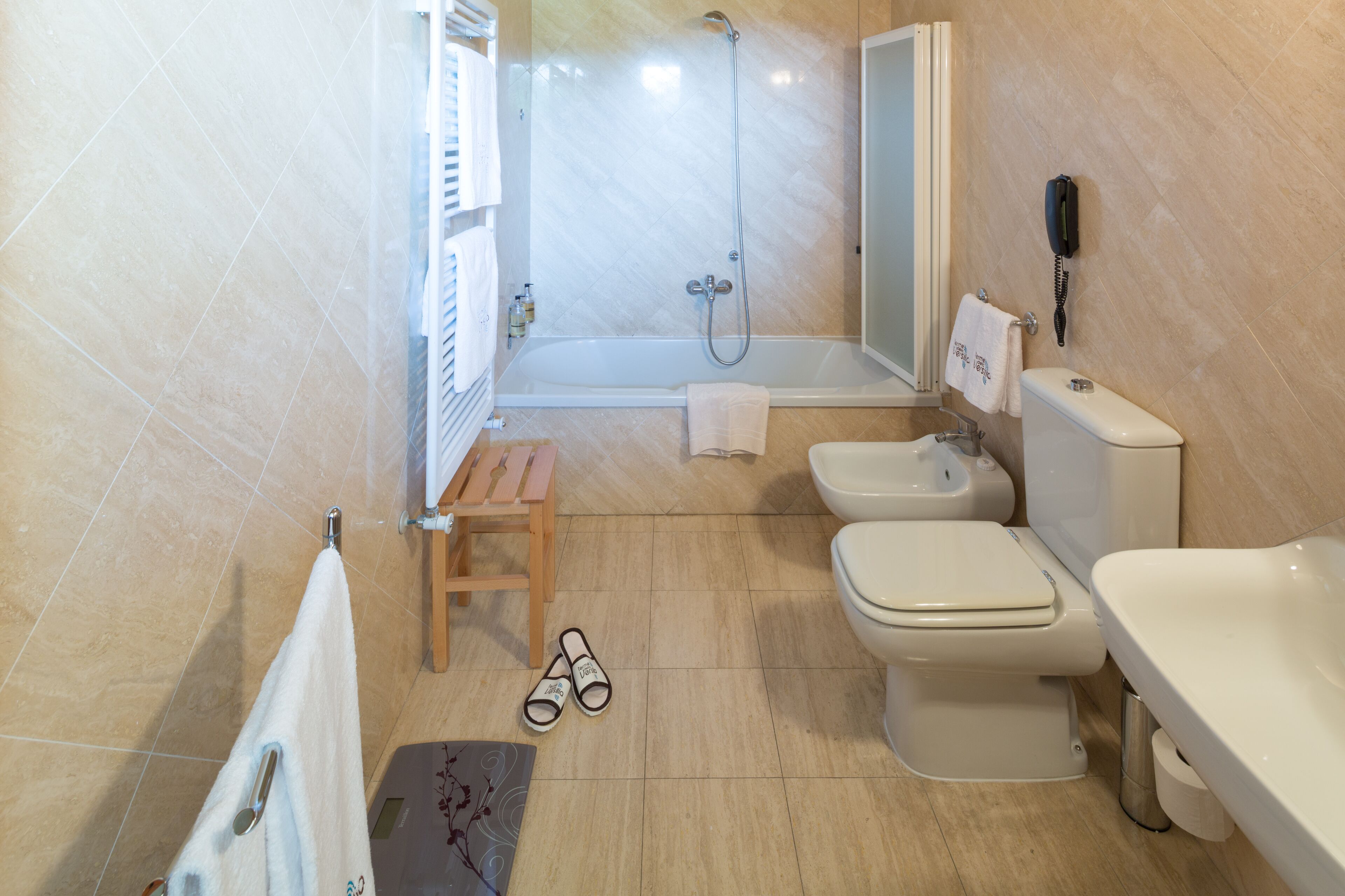 Un baño con bañera, inodoro, bidé y un taburete de madera.