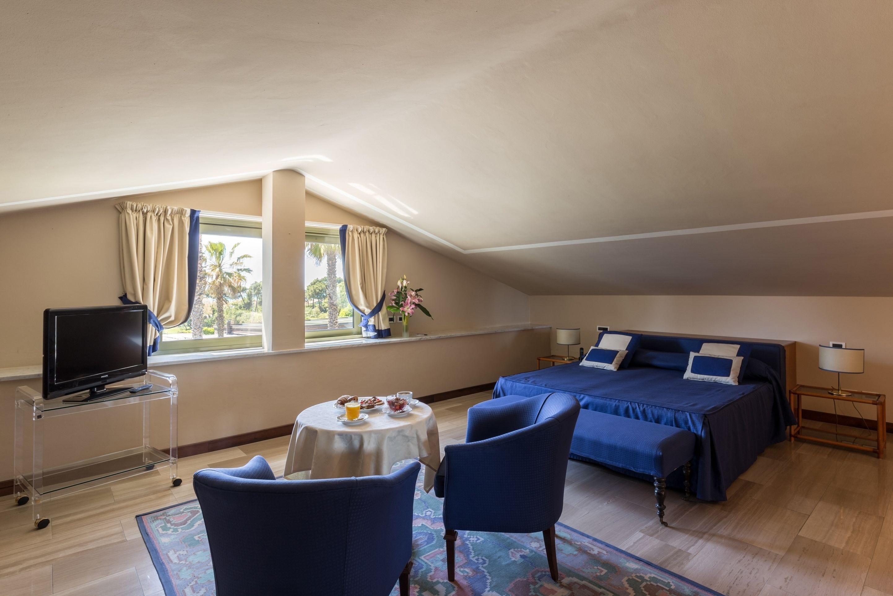 Une chambre à coucher avec un lit, deux chaises, une petite table avec une lampe, un téléviseur posé sur un meuble et une fenêtre avec des rideaux.