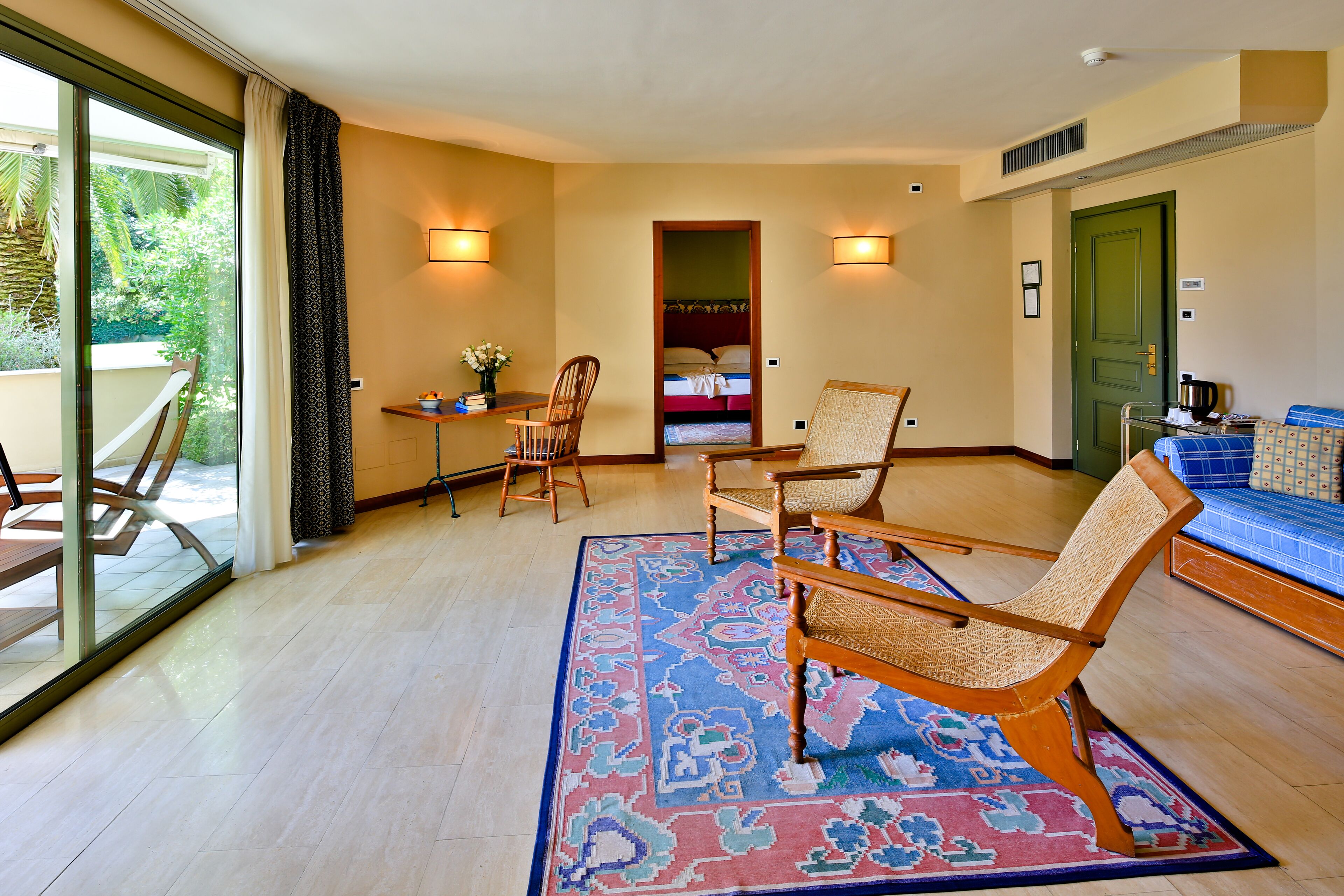 Camera d'hotel con un'ampia porta-finestra scorrevole che dà su un balcone, un letto con testata verde, due sedie di legno, un tavolino con un vaso e un tappeto blu decorato.
