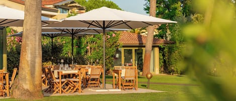 Un área de comedor al aire libre con mesas y sillas bajo sombrillas, rodeada de vegetación y un edificio al fondo.