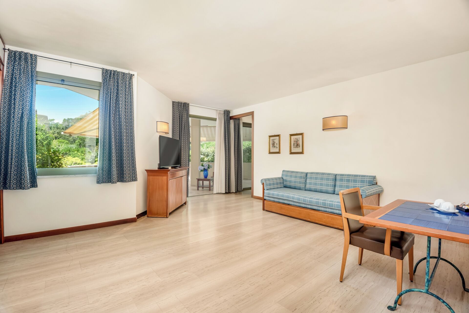 Una habitación con sofá, televisor sobre un mueble de madera, una mesa de comedor con sillas y vistas a zonas verdes al exterior.