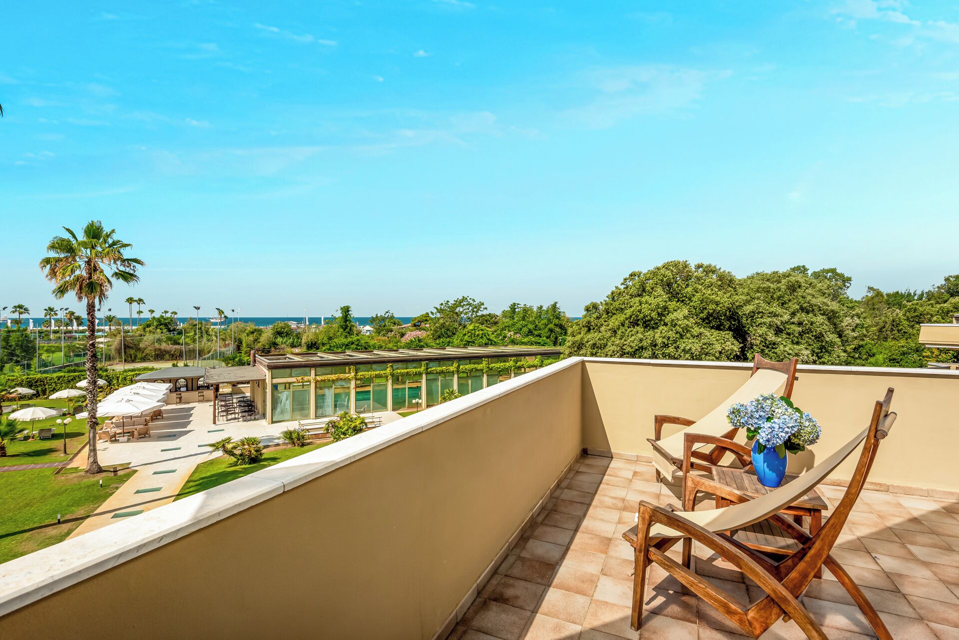 Un balcon avec vue sur une piscine et sur la verdure, des meubles en bois et un ciel dégagé.