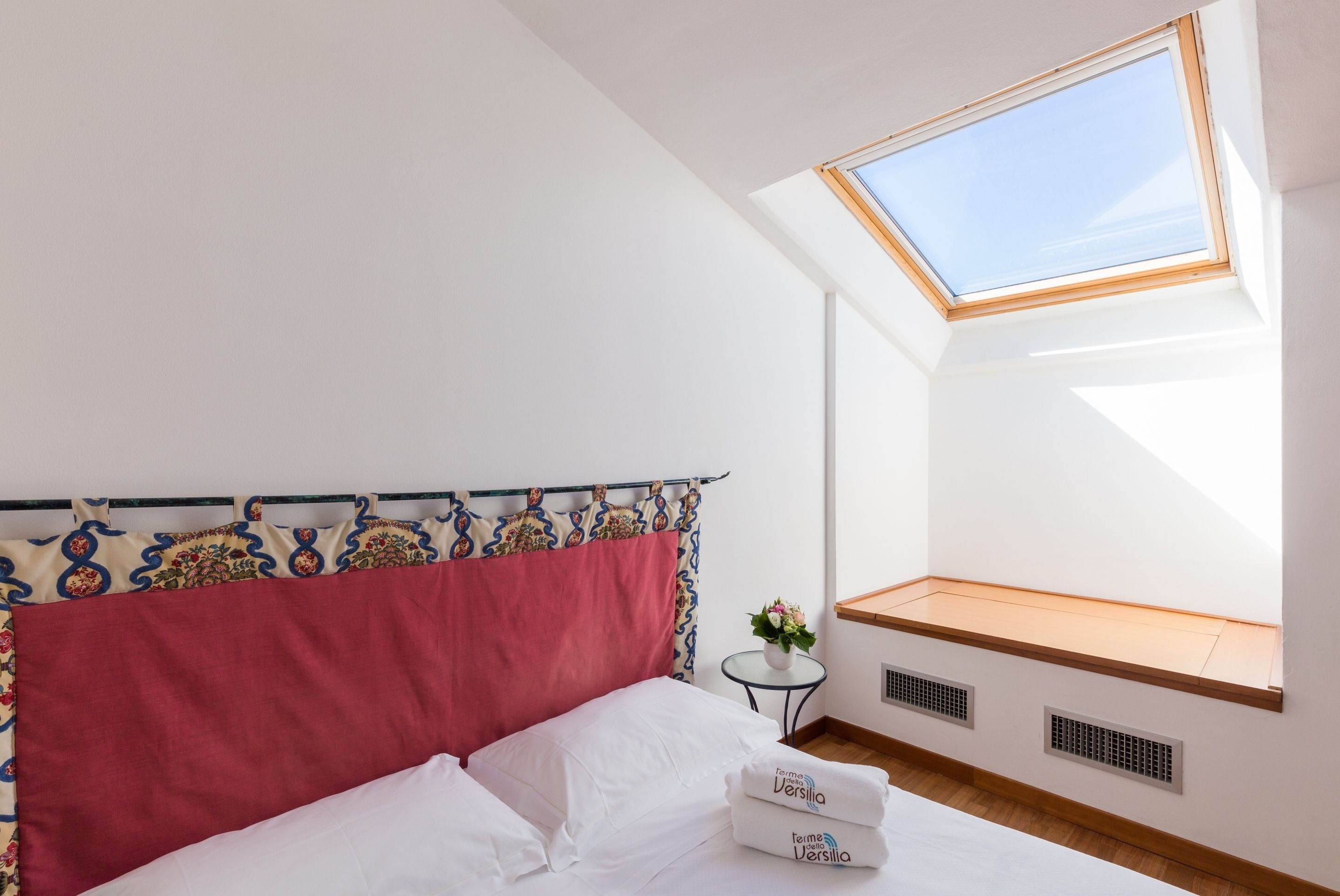 A bedroom with a skylight, a bed with white linens, a small table with a vase, and a window seat with a wooden ledge.