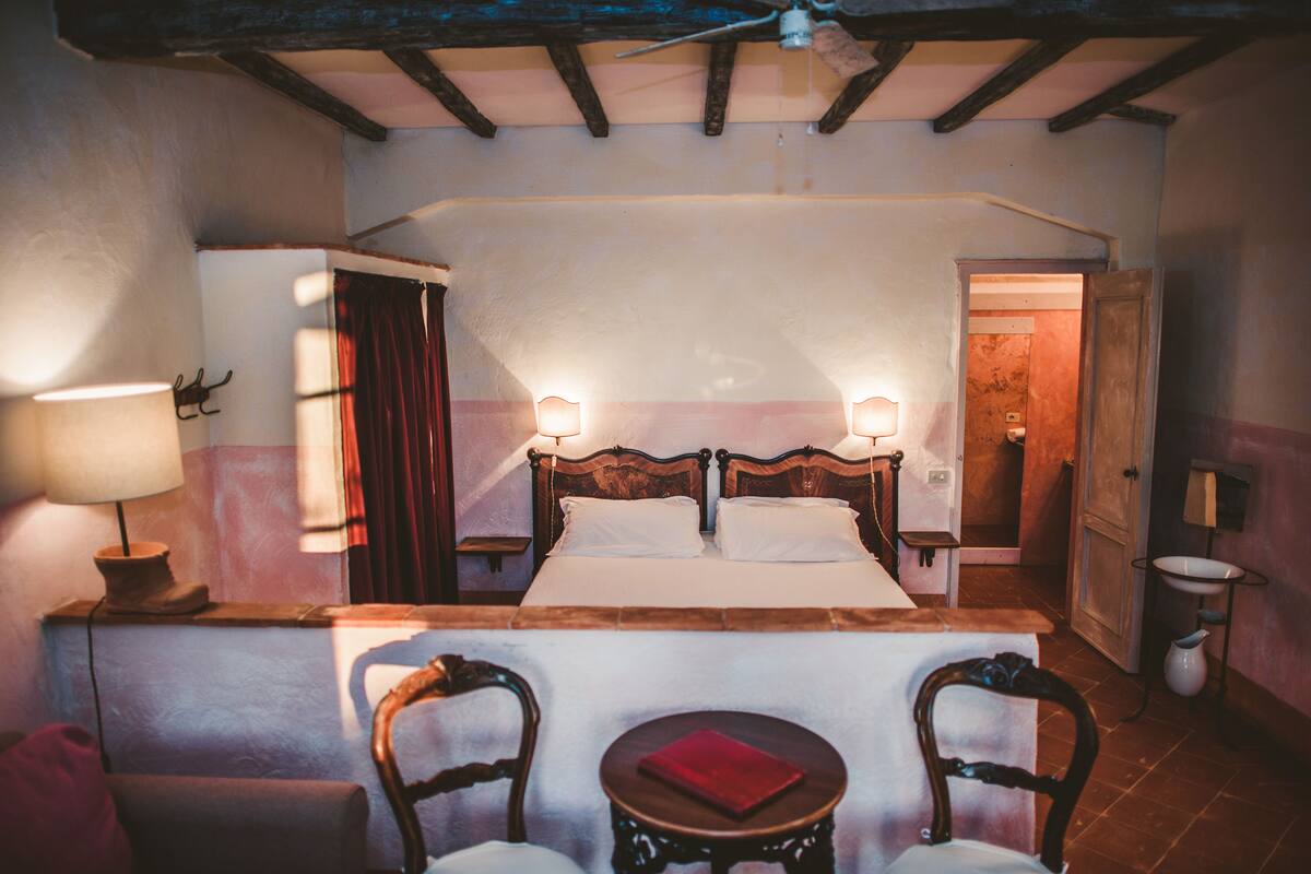 Ein Schlafzimmer mit einem hölzernen Bett, zwei Nachttischlampen, einem kleinen runden Tisch mit einem roten Buch und zwei Stühlen.