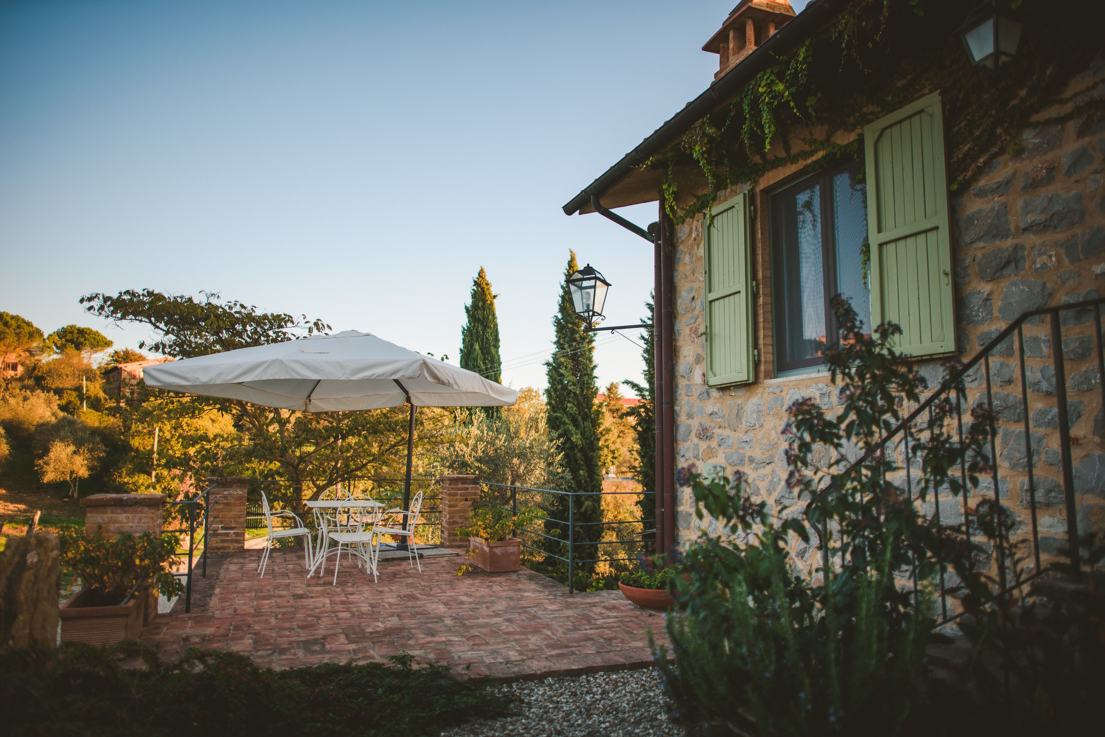 Une terrasse extérieure avec une table et des chaises sous un parasol blanc, entourée de plantes en pot et d’un mur de pierre.
