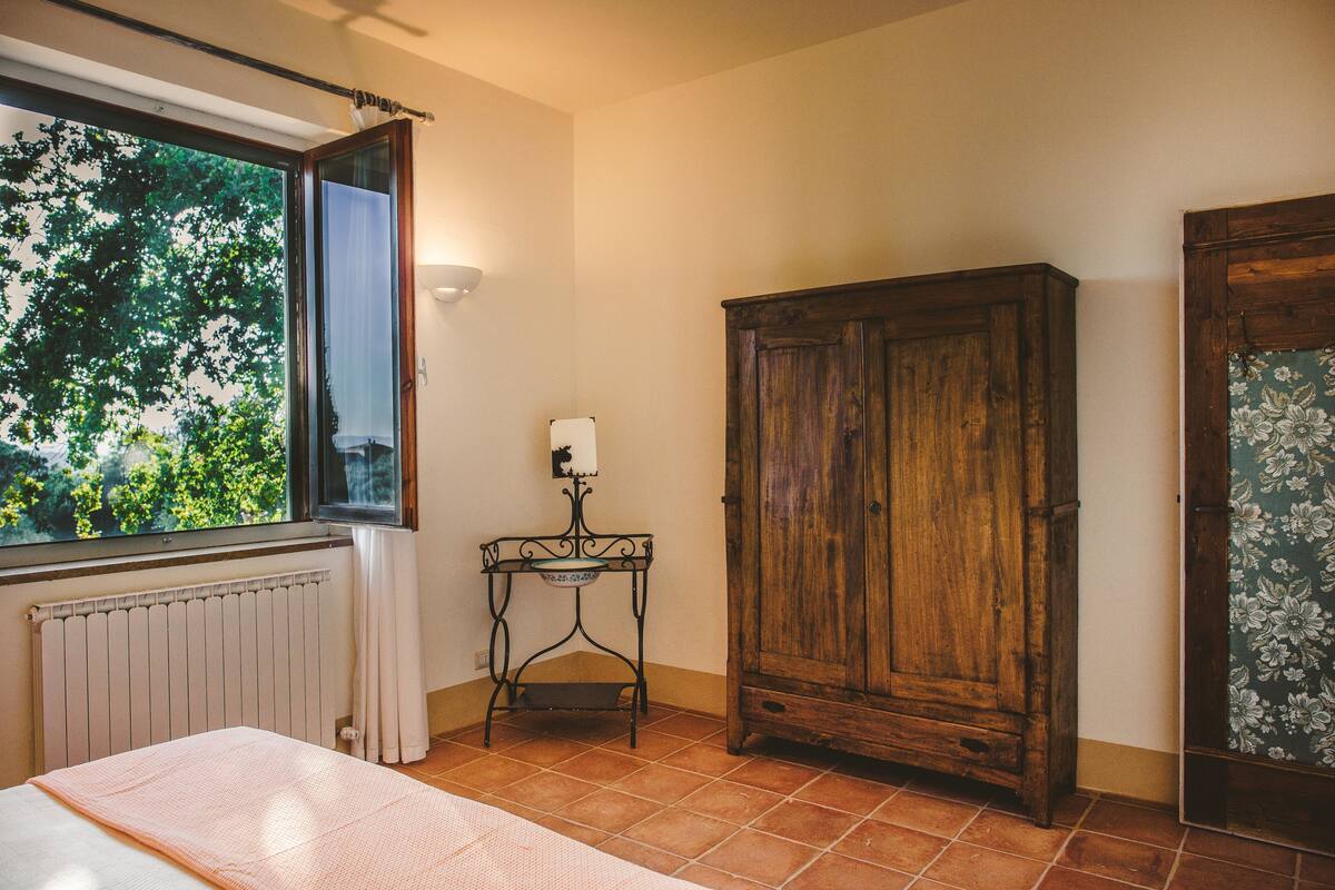Ein Schlafzimmer mit einem Bett, einem Fenster mit Vorhängen, einem Heizkörper, einem Holzschrank und einem kleinen Tisch mit einer Lampe.