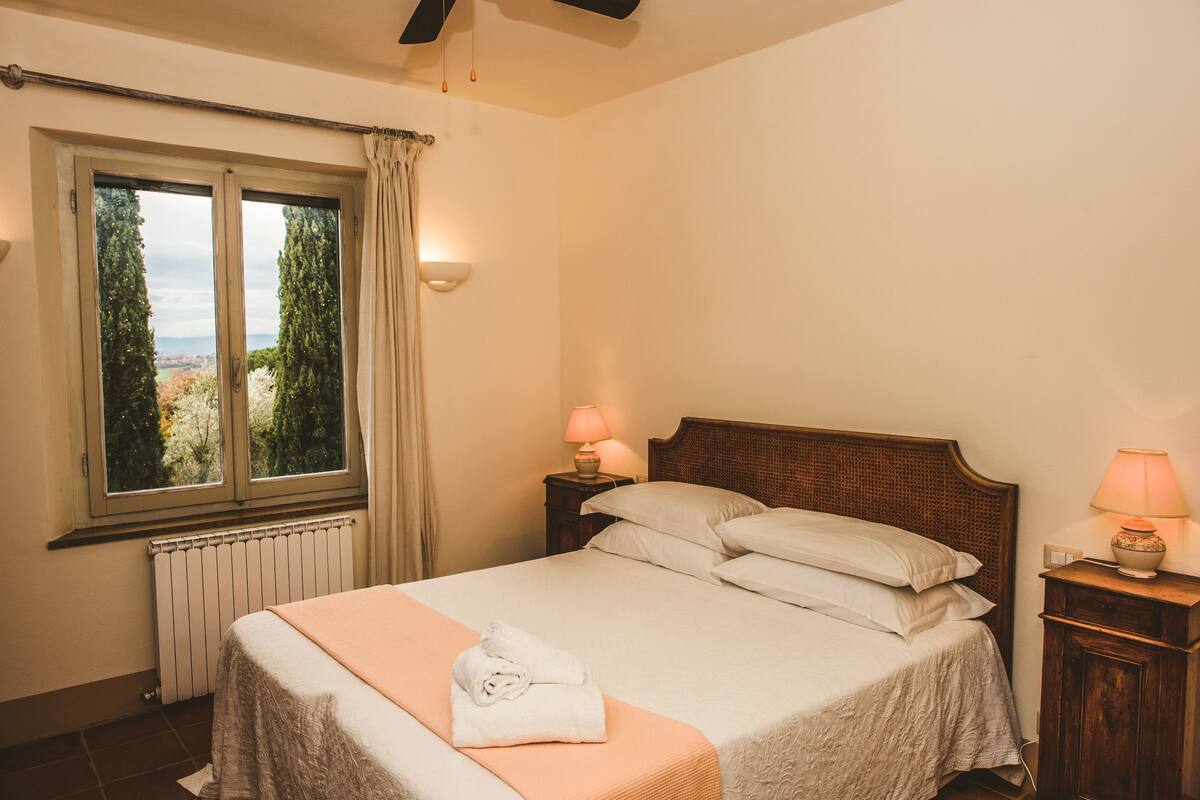 Ein Schlafzimmer mit einem Bett, zwei Nachttischlampen, einem Fenster mit Vorhängen und Blick auf Bäume.