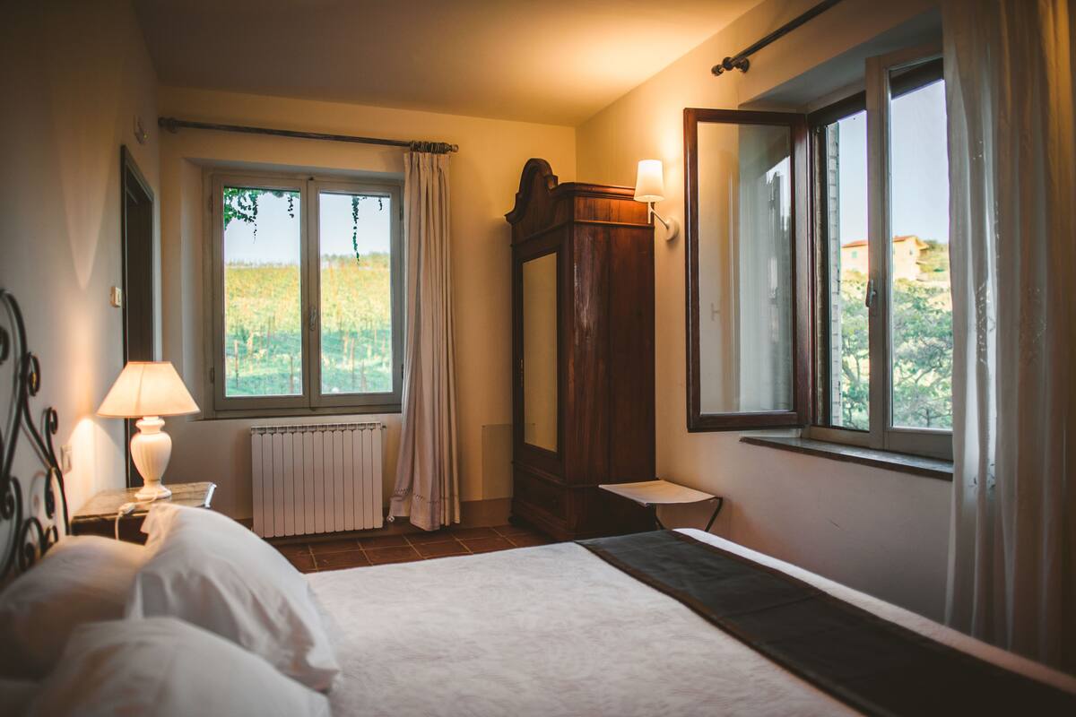 Ein Schlafzimmer mit Bett, Nachttisch, Lampe, einem Fenster mit Vorhängen, einer Heizung und einem Spiegel.