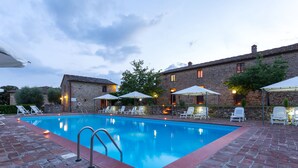 A swimming pool with lounge chairs and umbrellas, surrounded by stone buildings with outdoor lighting. - Tenuta di Monaciano (Castelnuovo Berardenga)