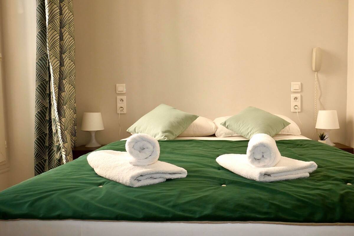 Cama bem arrumada com roupa de cama verde, dois toalhas enroladas e dois travesseiros, um verde e um branco.
