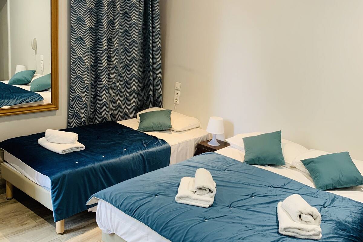 Quarto de hotel com duas camas, cada uma com colcha azul e travesseiros brancos. Há um espelho grande na parede, uma janela com cortinas e uma mesa de cabeceira com abajur.