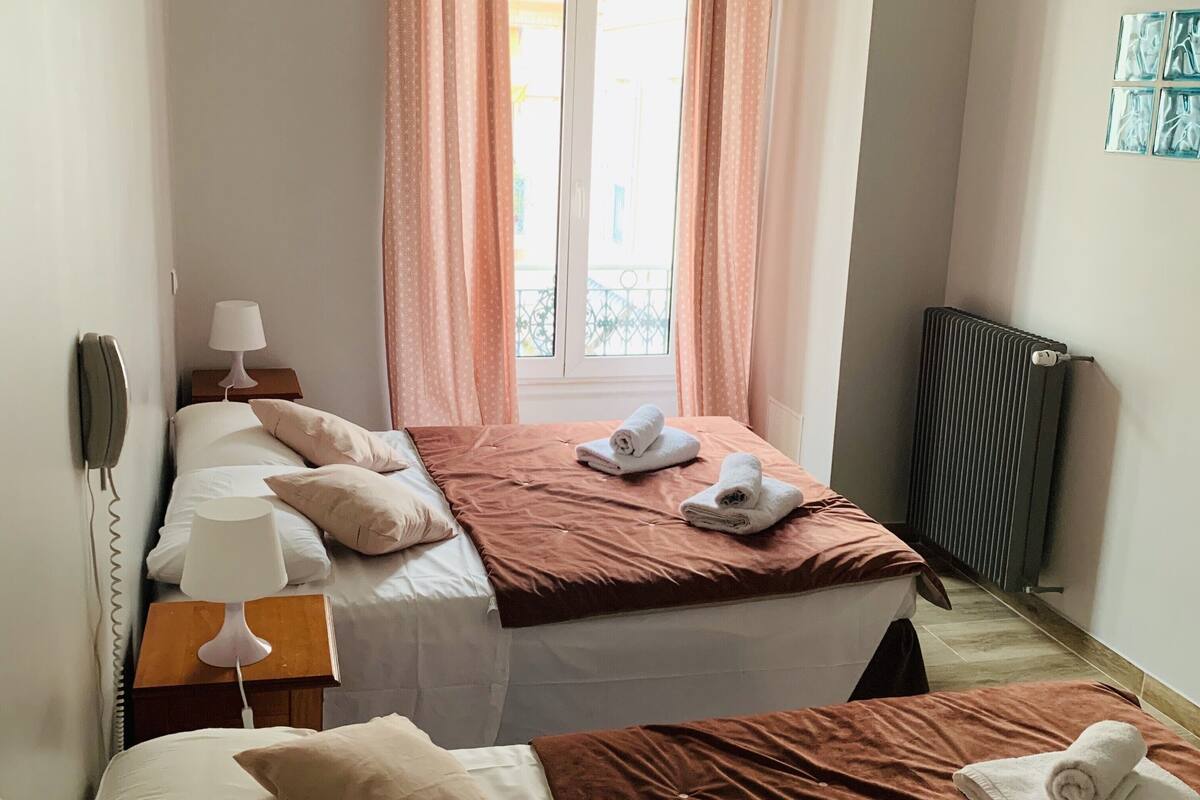Quarto de hotel com duas camas, um radiador, telefone e uma janela com cortinas.