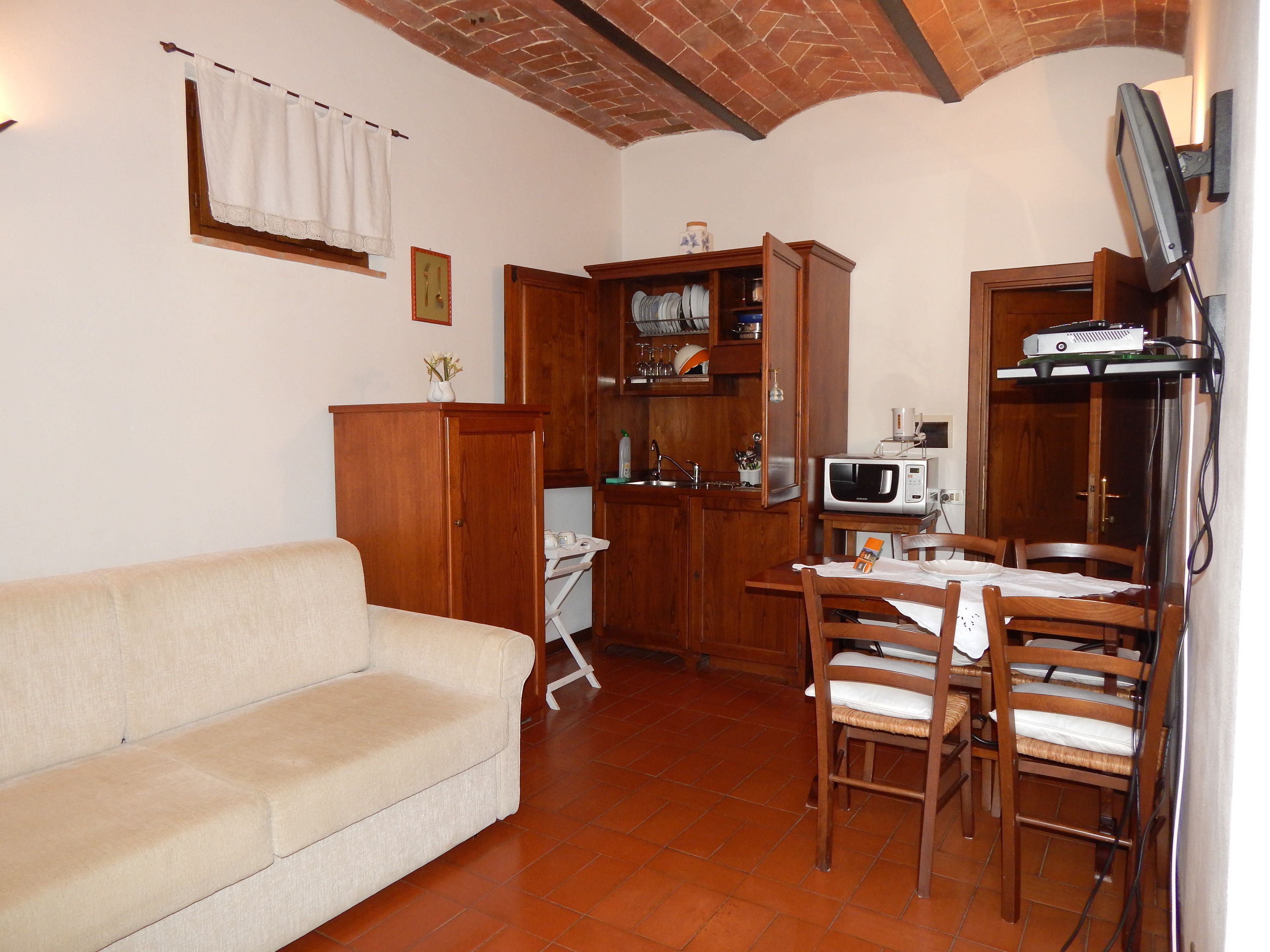 A compact living space with a sofa, wooden dining table, chairs, a kitchen area with cabinets and a microwave, and a brick ceiling.