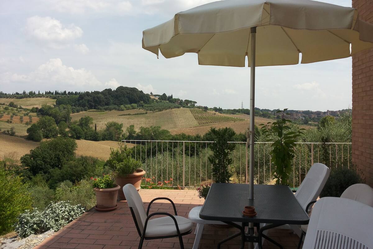 Un patio al aire libre con una mesa, sillas, un paraguas grande y una vista panorámica de colinas ondulantes y edificios lejanos.