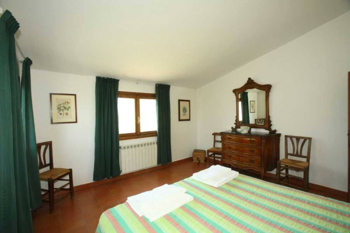 A bedroom with a bed, a dresser, two chairs, a window with curtains, and framed pictures on the walls.