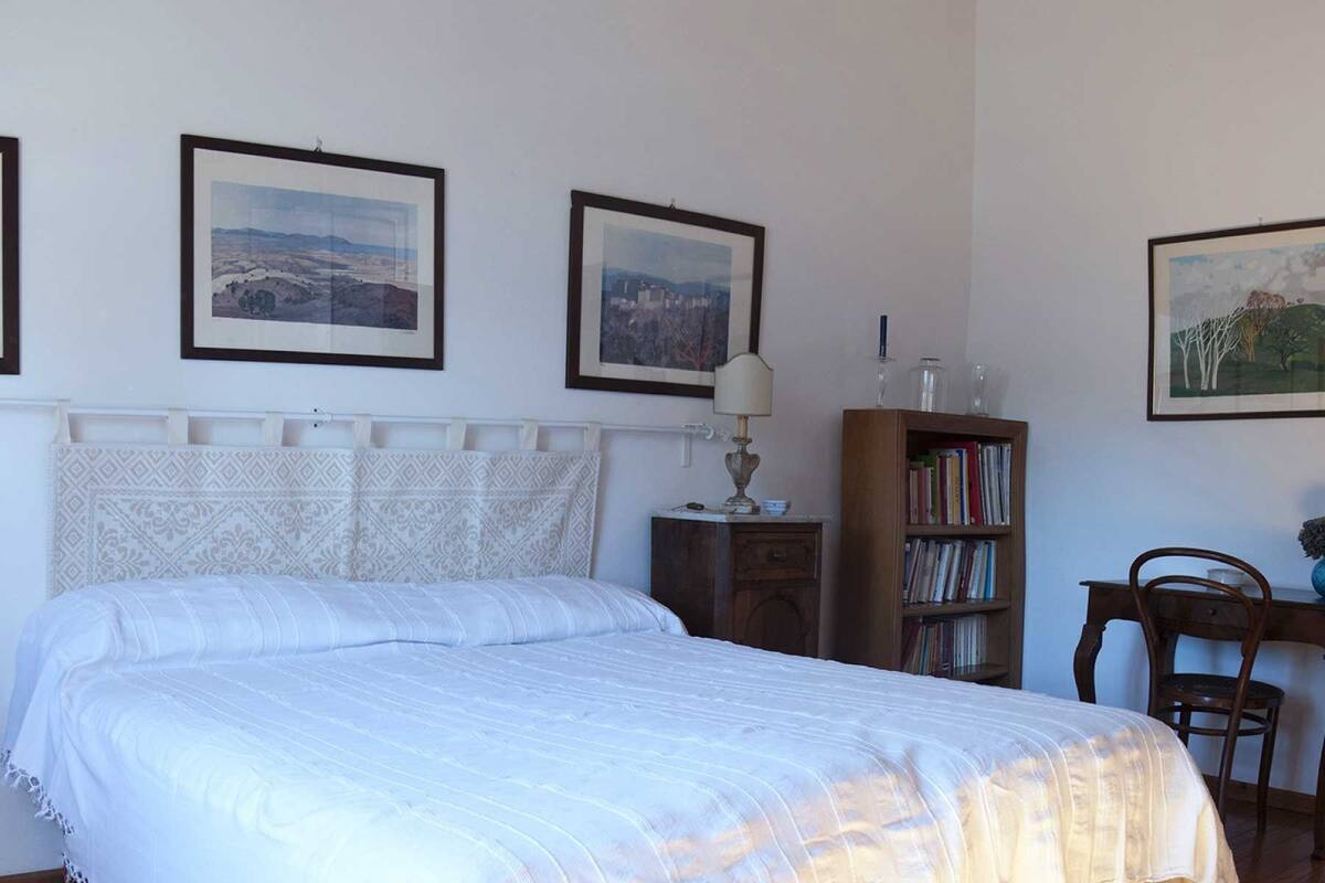A bedroom with a bed, a nightstand, a lamp, a bookshelf, and framed pictures on the wall.