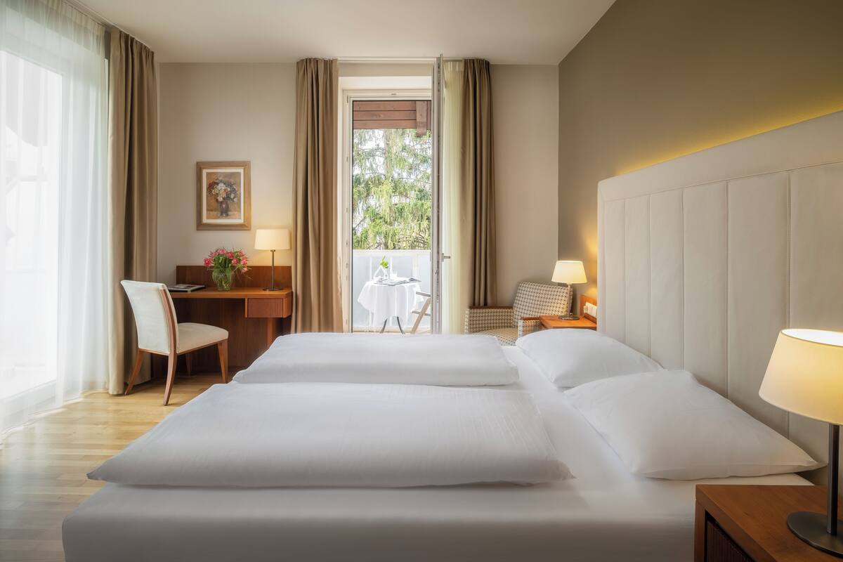 Quarto de hotel com duas camas, uma escrivaninha, uma cadeira, uma mesa pequena e vista para o exterior.