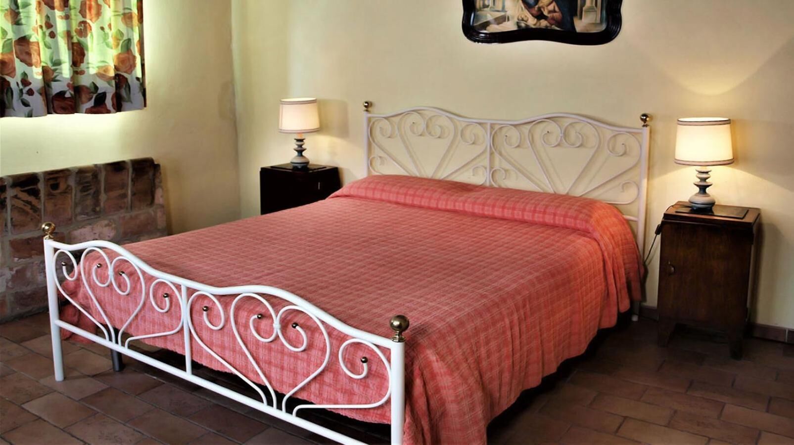 Un lit avec un cadre en métal blanc et une couette rouge, flanqué de deux tables de chevet avec des lampes, dans une pièce dont un mur est en briques et au-dessus du lit, on aperçoit un tableau encadré.