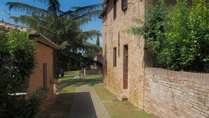 A narrow alley with a brick wall on one side and a building with a tiled roof on the other. There is a small garden area with a bench and a pathway leading to the building. - La Grotta di Montecchino (Siena)