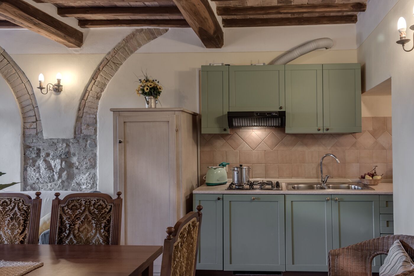 A kitchen with green cabinets, a stove, and a sink. There is a dining table with chairs and a decorative vase with flowers.