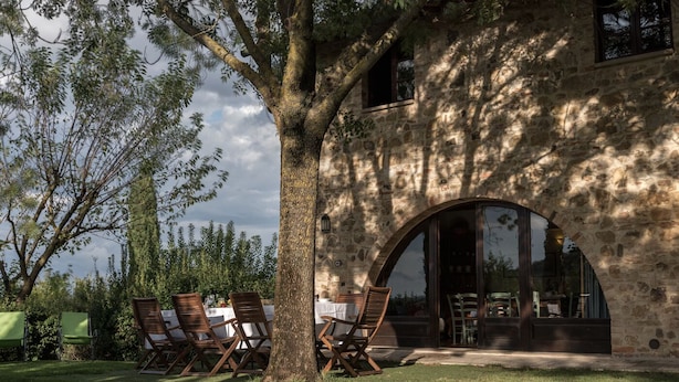 A stone building with a large tree in front, outdoor seating with tables and chairs, and a view of the surrounding landscape. - Borgo al Cerro (Casole d'Elsa)