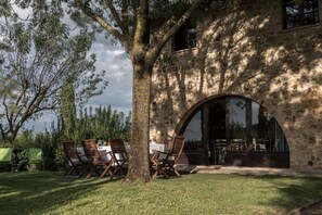 Un edificio in pietra con un grande albero di fronte, posti a sedere all'aperto con tavoli e sedie, e vista sul paesaggio circostante.