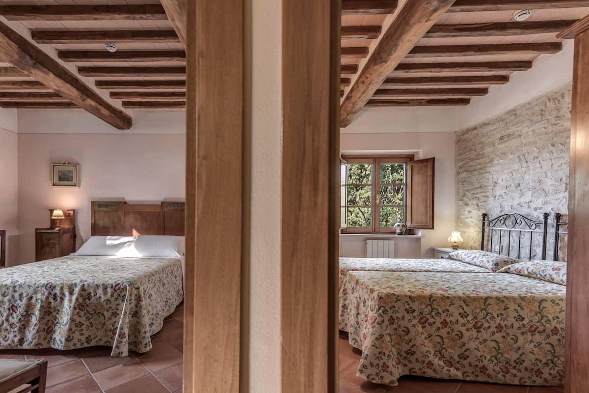 Une chambre avec deux lits, des poutres en bois au plafond, une fenêtre donnant sur un paysage verdoyant et un mur en pierre.