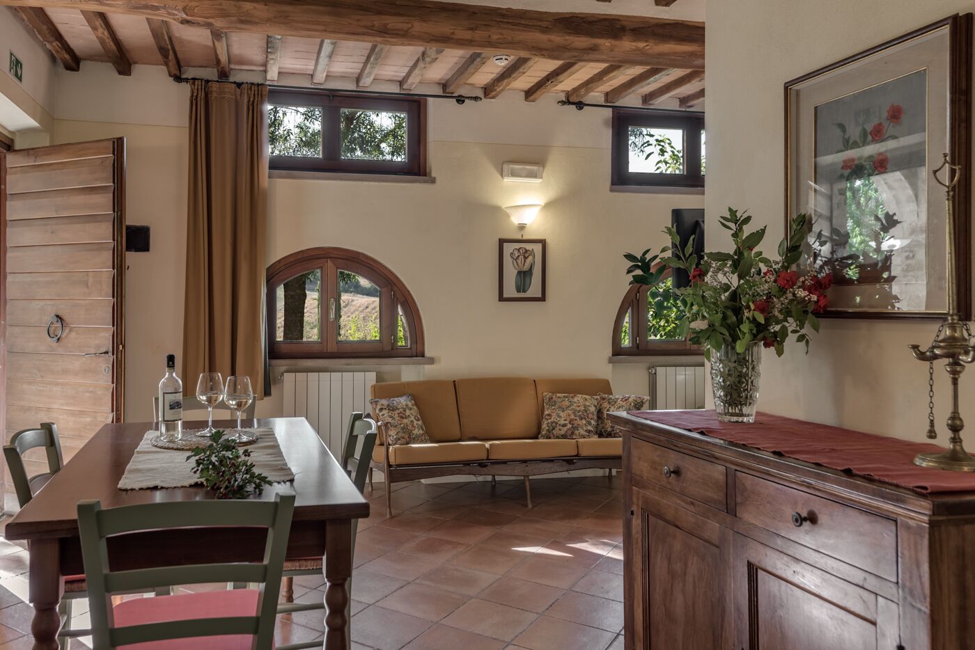 A dining area with a wooden table, chairs, and a sofa. There are windows with curtains, a radiator, and a vase with flowers on a cabinet.