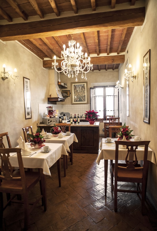 A dining area with wooden tables, chairs, a chandelier, and a kitchen in the background.