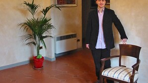 Un homme en costume, debout à côté d’une chaise dans une pièce ornée d’un lustre, d’une plante en pot et d’un tableau accroché au mur.