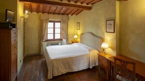 Une chambre à coucher comprenant un lit, des tables de chevet, une commode, une fenêtre avec des rideaux, un plafond avec des poutres apparentes et un mur orné de tableaux.