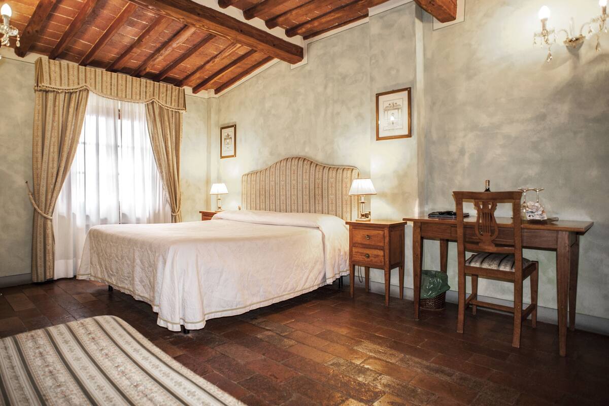 Quarto com teto de madeira, cama com colcha branca, duas mesas de cabeceira com luminárias, uma cadeira de madeira e uma escrivaninha com cadeira.