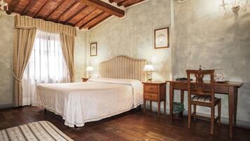 Quarto com teto de madeira, cama com colcha branca, duas mesas de cabeceira com luminárias, uma cadeira de madeira e uma escrivaninha com cadeira.