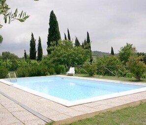Une piscine rectangulaire bordée d’une terrasse carrelée, entourée de verdure et de grands arbres.
