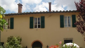 Un bâtiment doté d’un toit recouvert de tuiles, de volets verts et d’une entrée centrale surmontée d’un arc. Devant l’entrée, on trouve des plantes en pot ornées de fleurs rouges et roses.