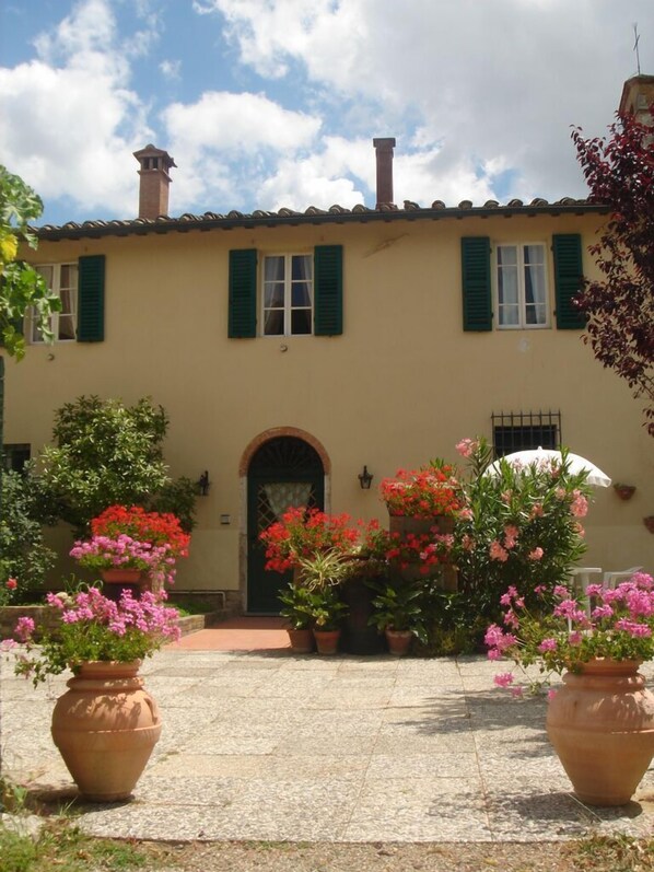 A building with a tiled roof, green shutters, and a central entrance with an archway. There are potted plants with red and pink flowers in front of the entrance.