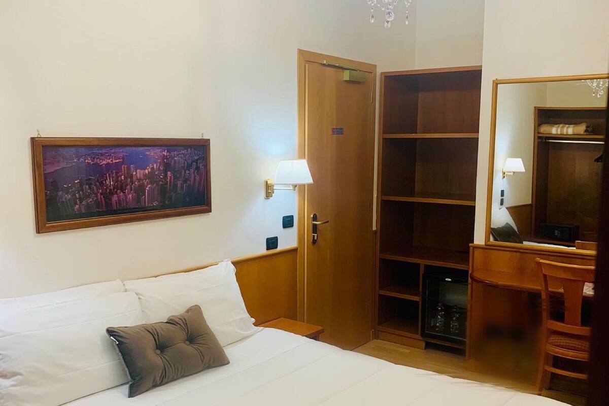 Una habitación de hotel con una cama grande, un armario de madera, un escritorio con silla, un espejo y una araña.
