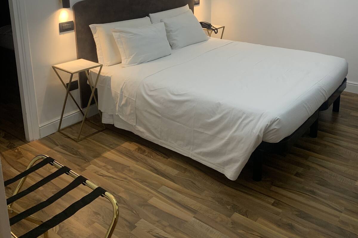 Una cama bien tendida con sábanas blancas, suelo de madera, aire acondicionado de pared y un espejo decorativo con pan de oro.