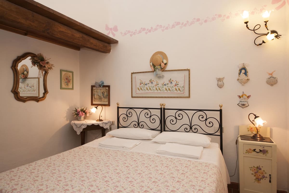 Una cama bien hecha con cubrecama floral, un cabecero de madera, una mesita de noche con lámpara, y adornos de pared que incluyen cuadros enmarcados y un aplique.