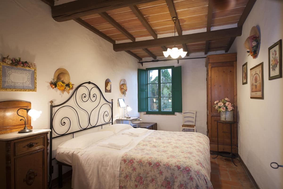 Un dormitorio con cama, cabecera de madera, mesita de noche con lámpara, cómoda y ventana con contraventanas verdes, además de una puerta de madera.
