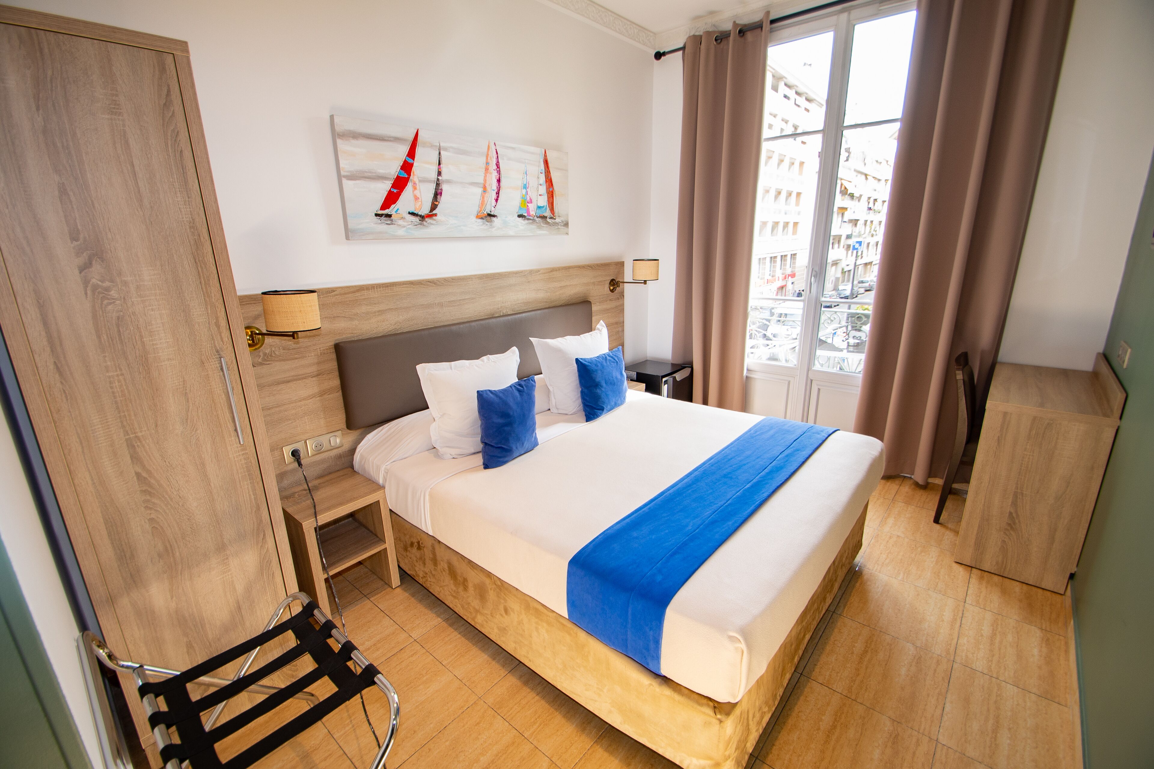 A hotel room with a large bed, a wooden wardrobe, a nightstand, a lamp, and a window with curtains.
