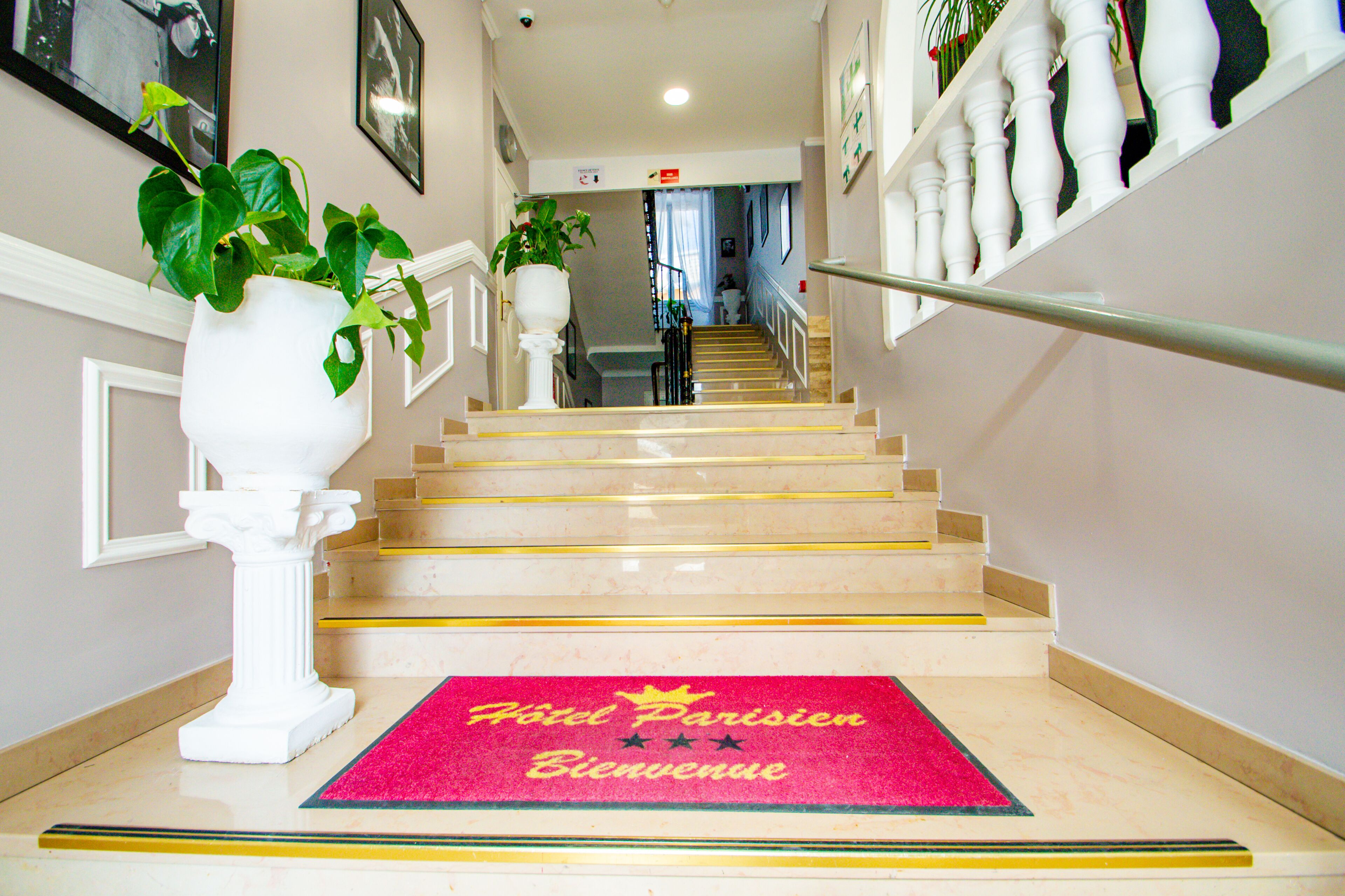 Un escalier d’hôtel recouvert d’un tapis rouge sur lequel on peut lire « Hôtel Parisien, bienvenue », et une colonne blanche ornée d’une plante.