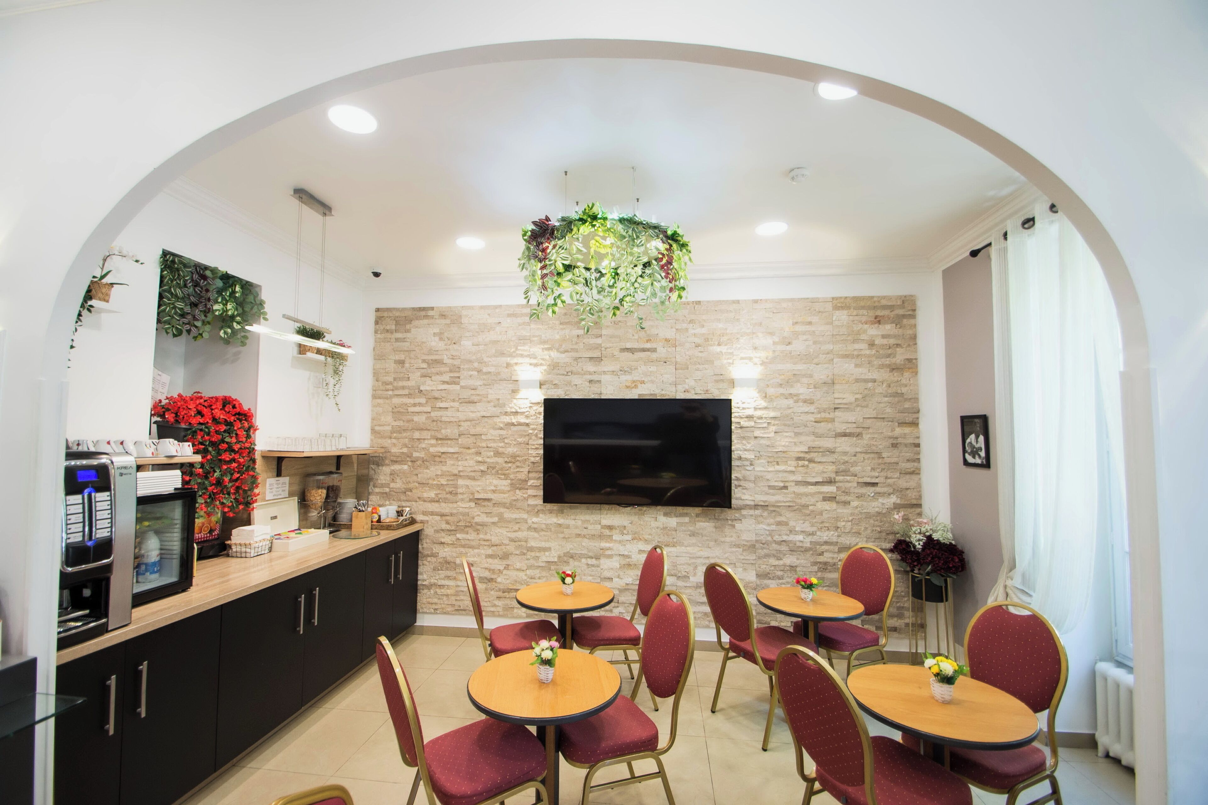 A modern kitchen with a stone backsplash, a flat-screen TV, and a dining area with red chairs and round tables.