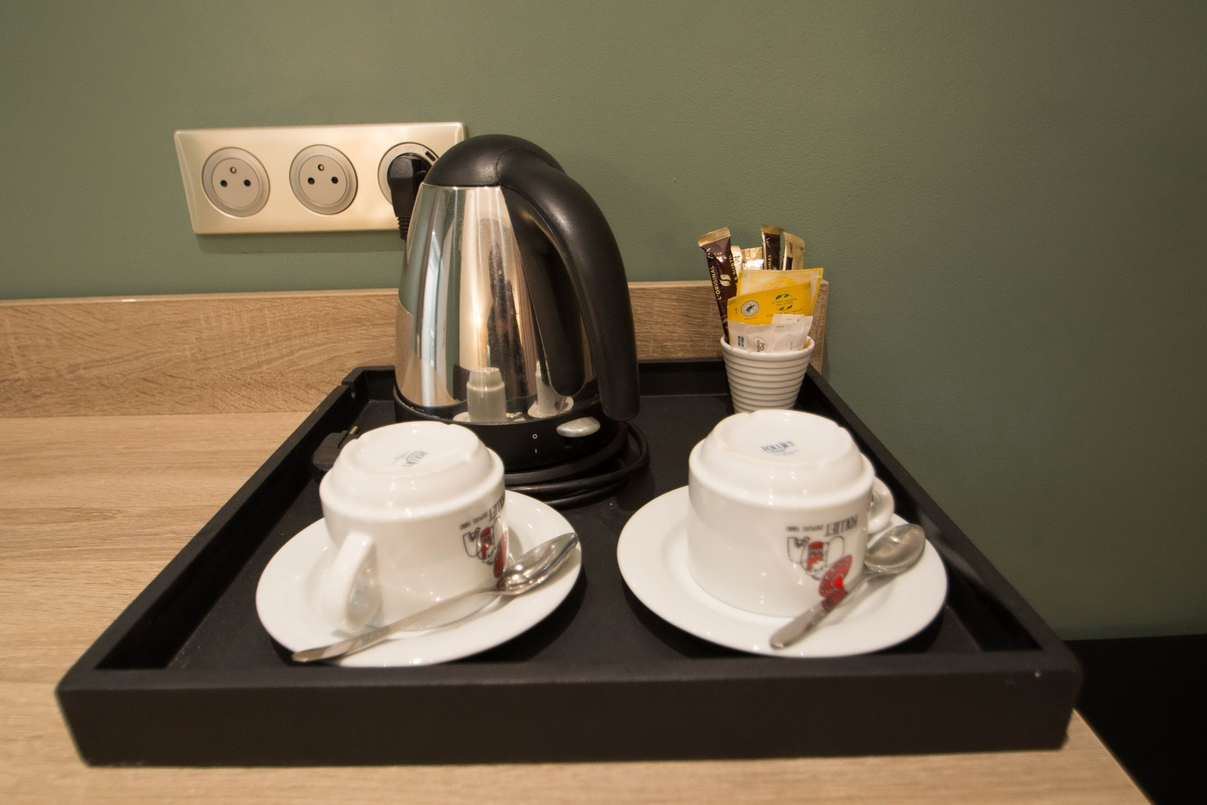A tray with two white cups and saucers, a black kettle, and a container with utensils.