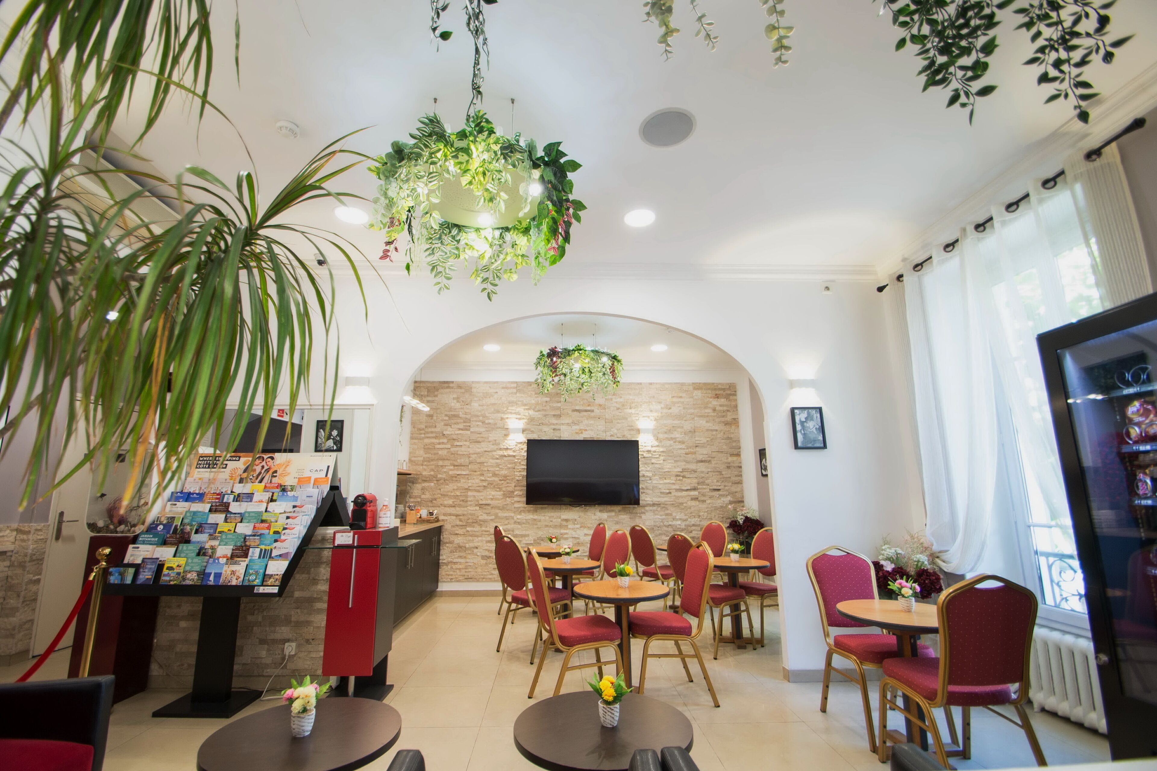 A hotel lobby with a reception desk, a wall-mounted TV, and a seating area with round tables and red chairs.