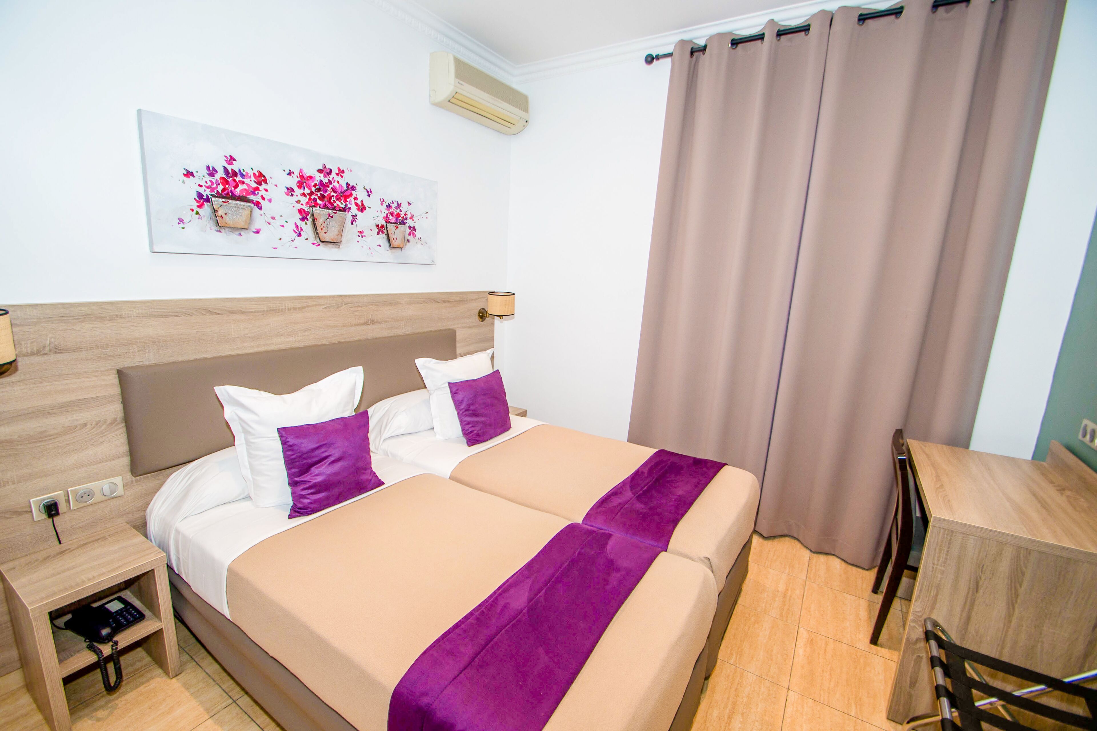 Een tweepersoonsbed met een houten hoofdbord, een nachtkastje met een telefoon, en een wandairconditioner.
